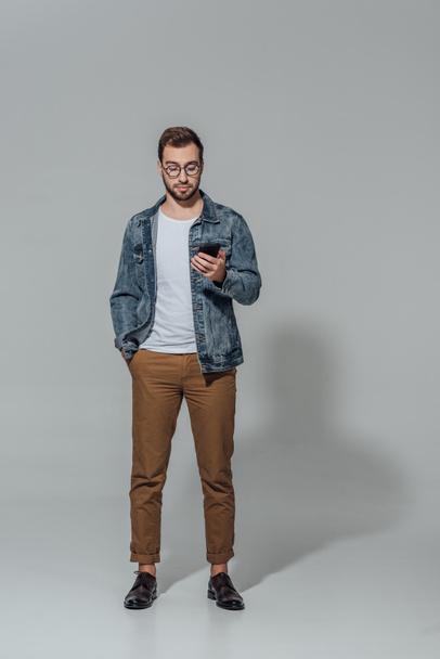 full length view of handsome sylish young man in eyeglasses using smartphone isolated on grey - Fotoğraf, Görsel