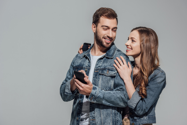 beautiful young couple in denim jackets using smartphones and smiling each other isolated on grey - Φωτογραφία, εικόνα