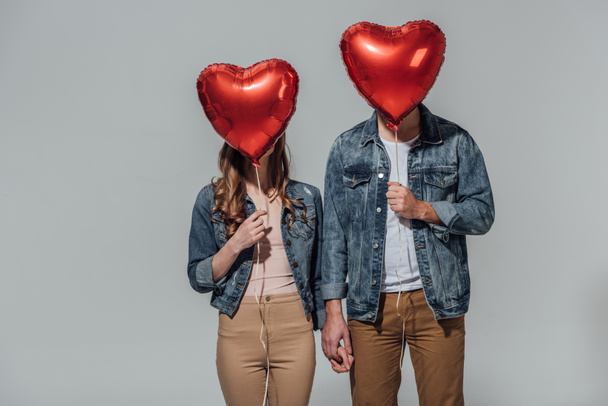νεαρό ζευγάρι κρύβεται πρόσωπα πίσω από την κόκκινη καρδιά σχήμα μπαλόνια που απομονώνονται σε γκρι - Φωτογραφία, εικόνα