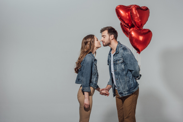 vue latérale du jeune couple capable de s'embrasser tandis que l'homme tenant des ballons rouges en forme de coeur isolé sur gris
  - Photo, image