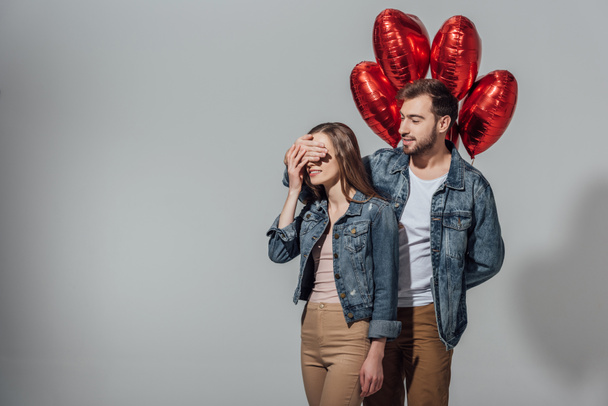 young man holding red heart shaped balloons and closing eyes to happy girlfriend isolated on grey - Photo, Image