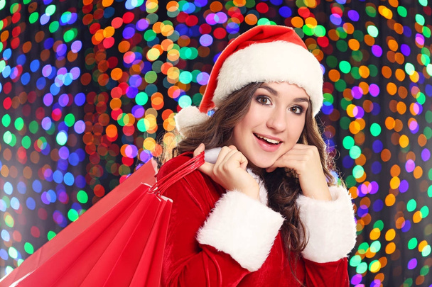 Young woman in santa hat with shopping bags on lights background - Fotoğraf, Görsel