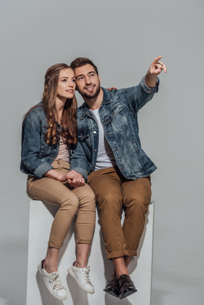 beautiful smiling young couple sitting together and pointing away with finger isolated on grey - Photo, Image