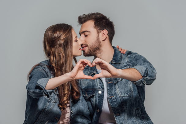 mooie jonge paar verliefd zoenen en tonen hand hartsymbool geïsoleerd op grijs - Foto, afbeelding