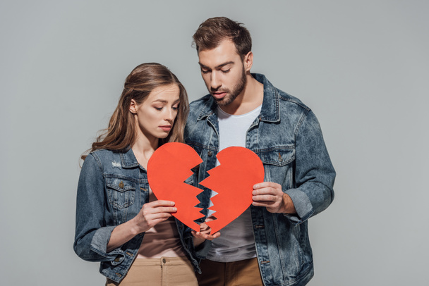 joven pareja molesta sosteniendo partes del símbolo del corazón roto aisladas en gris
  - Foto, Imagen