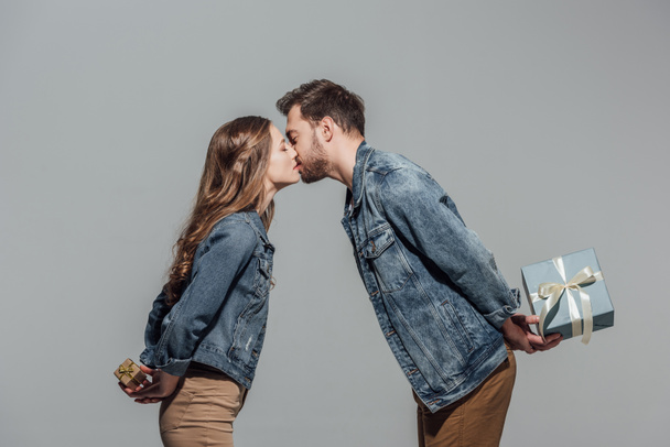 side view of beautiful young couple in love holding gift boxes and kissing isolated on grey - Photo, Image