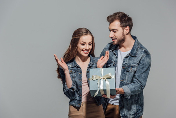 handsome young man presenting gift box to surprised girlfriend isolated on grey - Foto, immagini