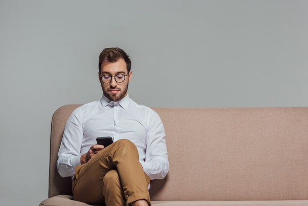 hansome joven en gafas con teléfono inteligente mientras está sentado en el sofá aislado en gris
 - Foto, Imagen