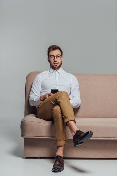 young man in eyeglasses holding smartphone and looking at camera while sitting on sofa isolated on grey - Zdjęcie, obraz