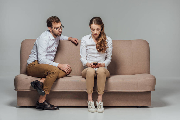 young woman using smartphone while boyfriend sitting near on sofa isolated on grey - Zdjęcie, obraz