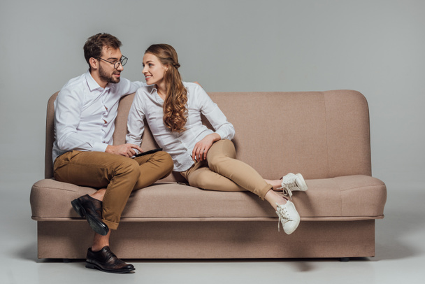beautiful smiling young couple using smartphone together while sitting on sofa isolated on grey  - Foto, afbeelding