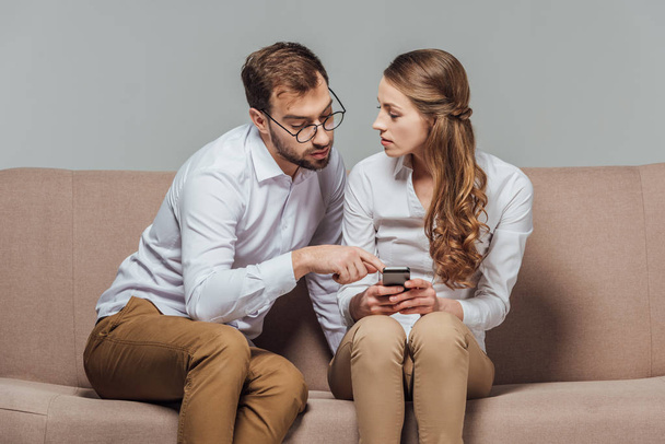young woman holding smartphone and handsome man in eyeglasses pointing with finger while sitting on sofa isolated on grey - Φωτογραφία, εικόνα