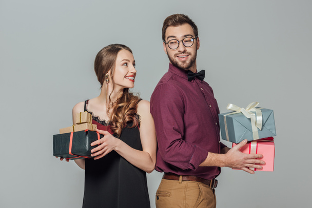 happy stylish young couple holding gift boxes and smiling each other isolated on grey - Photo, Image