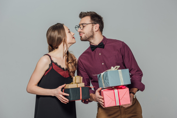 pareja joven feliz de moda sosteniendo cajas de regalo y capaz de besar aislado en gris
 - Foto, Imagen