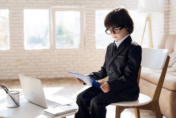 A little boy in a suit presents himself as a businessman. The dark-haired boy plays a rich man. He holds a folder and next to it stands a laptop on the desk. - Фото, изображение