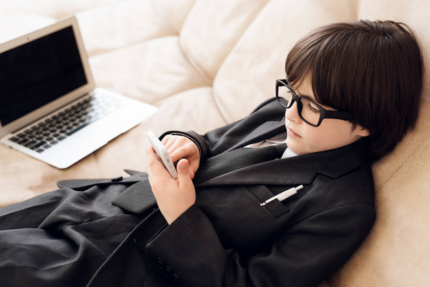 A little boy in a suit presents himself as a businessman. The dark-haired boy plays a rich man. A small businessman is lying on the couch and is looking at the phone. Next to it is a laptop. - 写真・画像