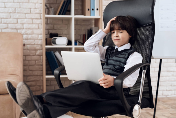 A little boy in a suit presents himself as a businessman. The dark-haired boy plays a rich man. A small businessman is sitting in a chair and working on a laptop. He is holding his head. - Photo, image