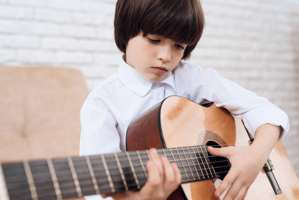A little dark-haired boy in a white shirt is playing the guitar. The boy learns playing the guitar himself. He sits on the couch in a bright room.x - Фото, изображение