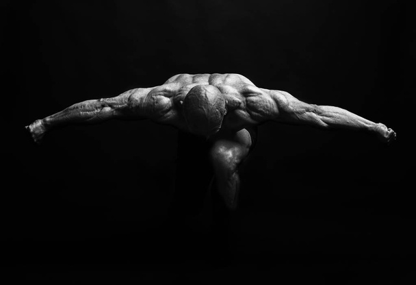 Μυώδης άνδρας με τα χέρια απλωμένα προς την πλευρά και το κεφάλι κάτω στο σκοτάδι - Φωτογραφία, εικόνα