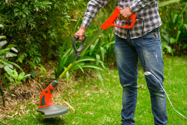 Young worker wearing jeans and long sleeve shirt and using a lawn trimmer mower cutting grass in a blurred nature background - Foto, Imagem