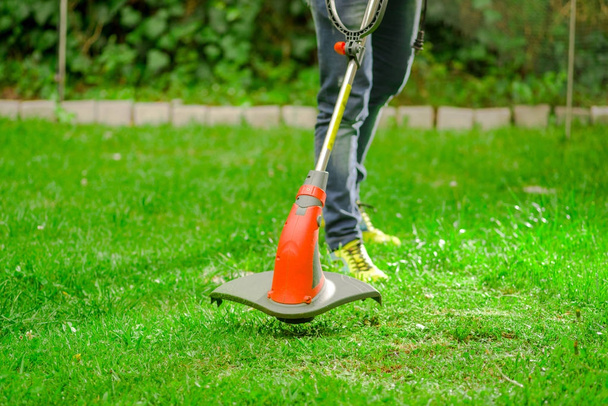 Outdoor view of young worker wearing jeans and using a lawn trimmer mower cutting grass in a blurred nature background - Foto, Imagen