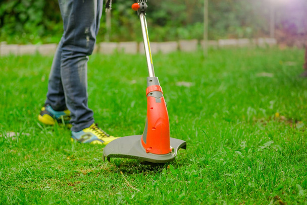 Young worker wearing jeans and using a lawn trimmer mower cutting grass in a blurred nature background - Foto, Imagen