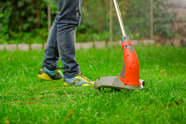 Close up of young worker wearing jeans and using a lawn trimmer mower cutting grass in a blurred nature background - Foto, Imagen
