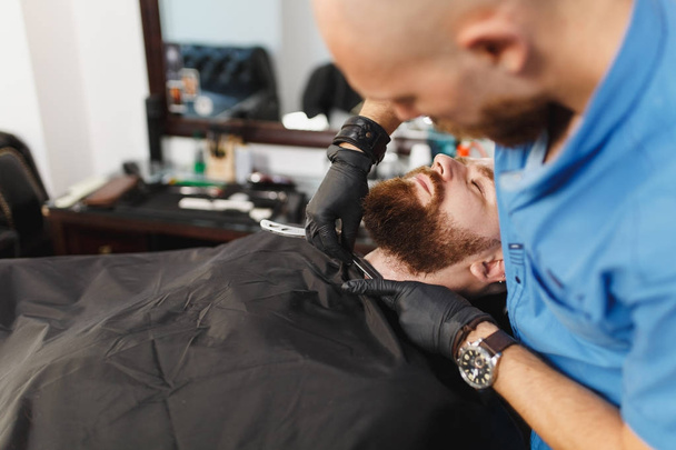 Male professional hairdresser serving client, shaving thick big beard straight razor. Ginger handsome stylish young man with short hair getting trendy haircut, black cape. Light white barber shop room - Foto, Imagem