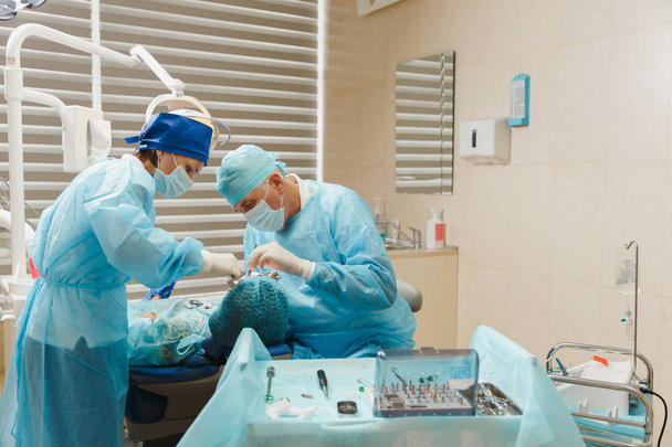Cirurgião dentista profissional velho masculino em uniforme e assistente feminino ajuda a realizar a operação instalar dentes de implante dentário de paciente mulher no escritório de luz clínica com equipamento de ferramentas modernas
. - Foto, Imagem