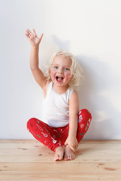 Photo of joyful blonde small kid sits on wooden floor, dressed casually, raises hand, isolated over white background. Cute lovely girl with cheerful expression. Children and emotions concept. - Foto, immagini