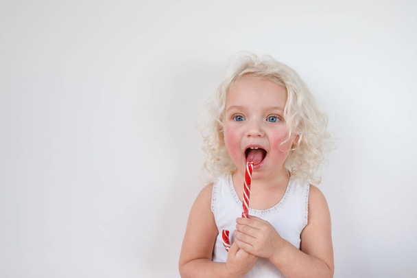 Horizontal shot of attractive blue eyed female kid has rough cheeks, looks thoughtfully aside, licks red and white sweet caramel cane, isolated over white background. Lovely small child indoor - Фото, изображение