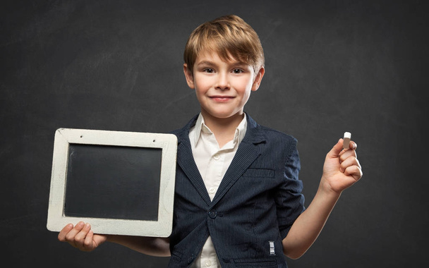 Handsome 7 year old School Boy holding a Cute little blackboard with Copy Space against a Dark Black Board Background: Confident and happy - Zdjęcie, obraz