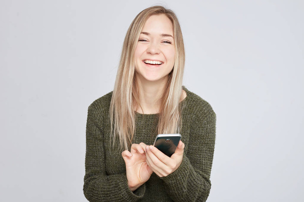 Studio shot of pretty young laughing European woman, showing perfect tooth smile, typing message to boyfriend on mobile phone, holding gadget with one hand, wearing warm wool sweater, looking happy. - Photo, Image