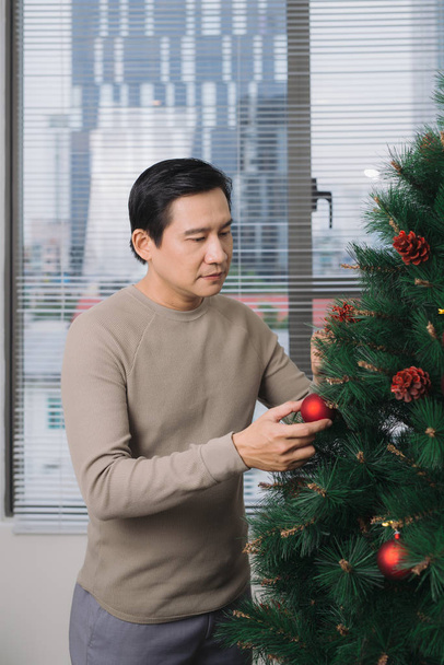 Celebrating New Year. Asian man standing by new christmas tree at home  - Photo, Image