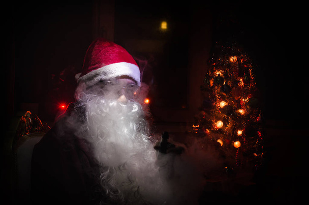 Papai Noel vaping cigarro eletrônico vestido de Papai Noel tradicional em um fundo tonificado escuro com nuvens de vape. Foco seletivo
 - Foto, Imagem