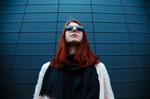 Hipster redhaired girl in stylish sunglasses is posing in front of a black wall on the street - Foto, Imagen