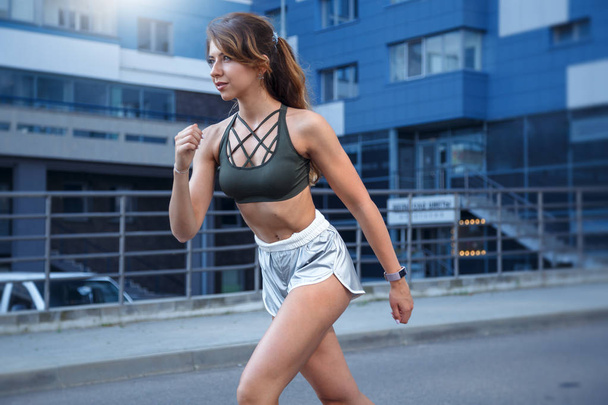 Long haired sporty girl in a stylish silver colored shorts and dark green t-shirt is exercising on a urban background.. The photo is made durring the sunrise in a lovely summer morning - Foto, Bild