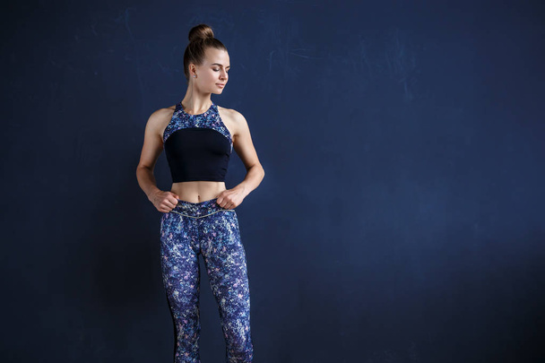 Beautiful fitness model is posing in front of the dark wall in a dark training suit - Zdjęcie, obraz