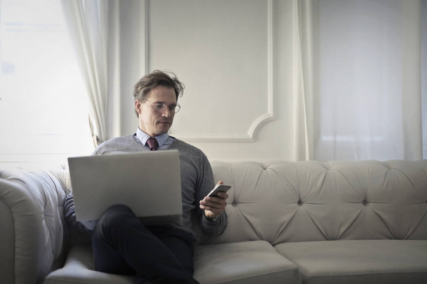 Elegant man using a laptop and a smartphone sitting on a sofa - Fotografie, Obrázek