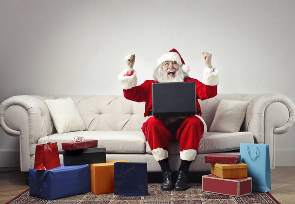 Santa Claus sitting on an armchair with a laptop on his knees exulting surrounded by gifs - Zdjęcie, obraz