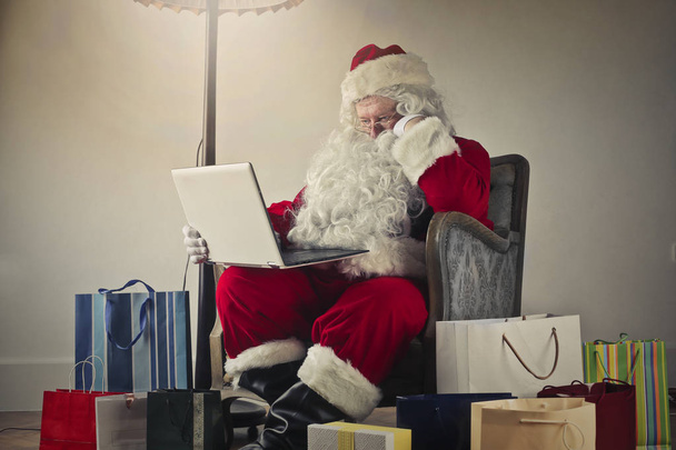 Santa Claus sitting on an armchair with a laptop on his knees surrounded by gifs - Foto, immagini
