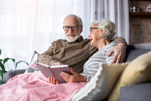 Delighted old couple sitting on sofa with book - Фото, изображение