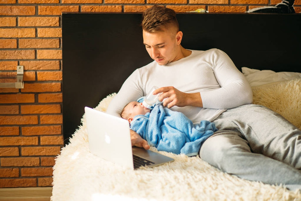 hombre alimentando pequeño bebé mientras está acostado en la cama y trabajando en el ordenador portátil
 - Foto, Imagen