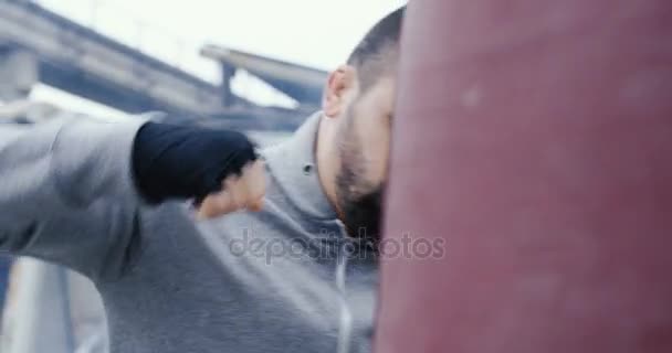 Close up of the handsome sportsman with a beard punching a red boxing bag on the abandoned factory area on the cold grey morning. Outdoors - Video, Çekim