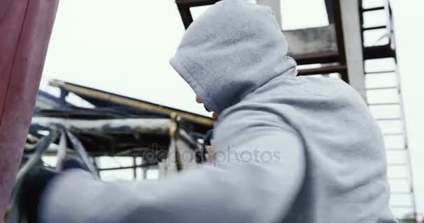 Rear of the close up of the handsome man with in a hood having boxing training with a red bag in the bad cold grey weather. Outside the big plant ruins. - 映像、動画
