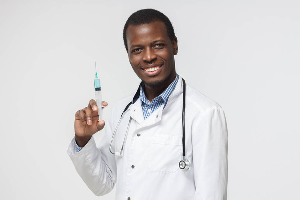Horizontal shot of young African doctor pictured isolated on white background wearing uniform holding syringe up getting ready for vaccination, smiling confidently, looking professional and positive - Photo, Image