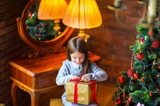 Christmas morning, beautiful little girl opens a gift sitting near the Christmas tre - Foto, Imagen