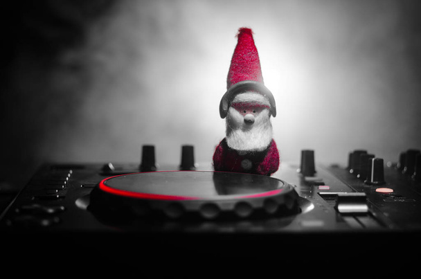 Mezclador de DJ con auriculares en el fondo oscuro club nocturno con árbol de Navidad Nochevieja. Vista de cerca de los elementos o símbolos de Año Nuevo (Santa Claus, muñeco de nieve, perro 2018, caja de regalo) en una mesa de Dj
. - Foto, Imagen