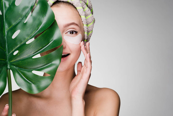 happy young girl with a towel on her head looks after the skin on her face, under the eyes of a patch, holds a green leaf - Foto, Bild