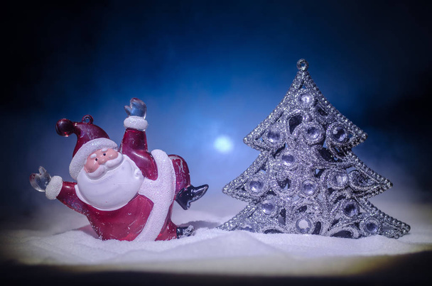 Ευτυχισμένος Βασίλη κούκλα την περίοδο των Χριστουγέννων με το δέντρο και το χιόνι. Bokeh πολύχρωμο φόντο. Santa Clause και καλά Χριστούγεννα παιχνίδι φιγούρα μοντέλο σε σκούρο φόντο τονισμένο ομίχλη. - Φωτογραφία, εικόνα